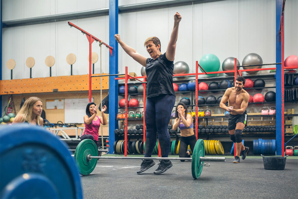 Crossfite - radujúca sa žena po cviku s veškou činkou, ľudia okolo jej tlieskajú